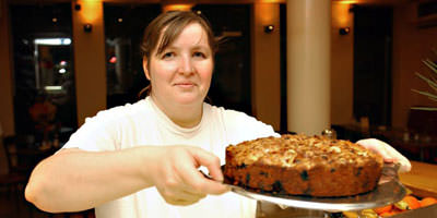 Foto: Eine Mitarbeiterin reicht einen leckeren Kuchen &uuml;ber die Glastheke. Er sieht noch warm aus mit braunen knusprigen Streuseln. Wahrscheinlich ist es ein Apfelkuchen, aber das beliebt eine Vermutung. 