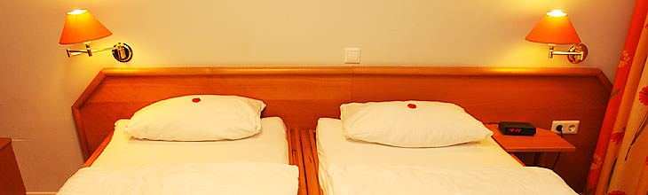 Panoramabild: Ein Doppelzimmer in warmer Atmosphäre. Zwei Leuchter tauchen es in sanftes, ruhiges Licht. Hinter den kleinen Marzipanherzen, die für jeden Besucher auf dem Kopfkissen liegen, spürt man die Menschen die dieses Hotel ausmachen.