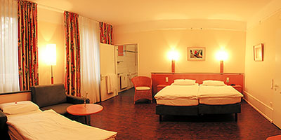 Panoramabild: Das grosse Familienzimmer in warmer Atmosphäre. Drei Leuchter tauchen es in sanftes, ruhiges Licht. Das Zimmer ist sehr geräumig und läßt am linken Rand einen Blick auf das barrierfrei Bad erhaschen.